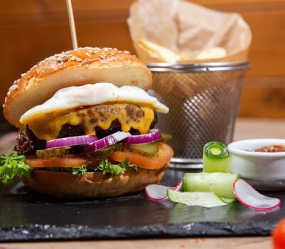 Burger cu ou, brânză, roșii, salată verde, ceapă și murături, alături de cartofi prăjiți și sos, pe o tablă neagră.