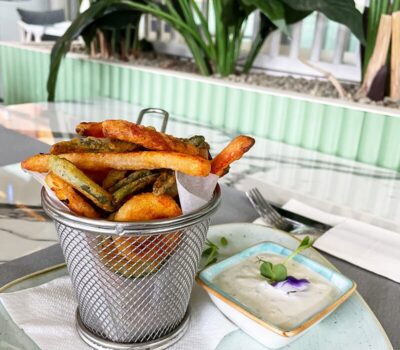 Un coș mic de metal umplut cu cartofi prăjiți și fasole verde și o farfurie cu sos de iaurt și o floare.