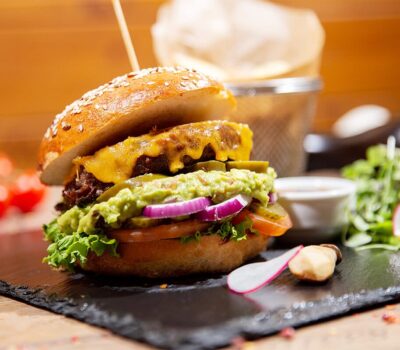 Un burger cu carne de vită cu brânză, guacamole, ceapă roșie, murături, roșii și salată verde pe o farfurie.