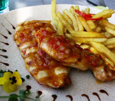 Două bucăți de pui la grătar cu sos și cartofi prăjiți pe o farfurie, cu flori galbene.