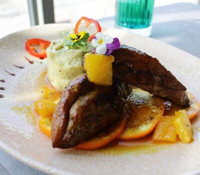 Carne de rață, piure de cartofi și felii de portocale pe o farfurie cu decorațiuni florale, cu o băutură în fundal.