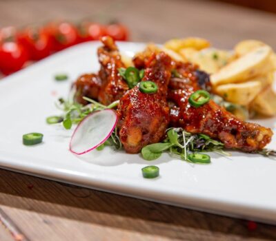 Aripioare de pui cu cartofi și garnitură, pe farfurie albă, cu roșii cherry în fundal.