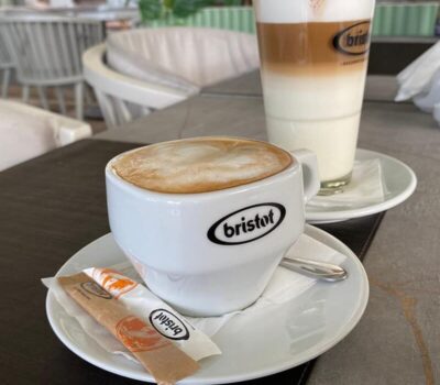 Cafea Bristot într-o ceașcă, zahăr, lingură și un pahar înalt cu pai dungat.