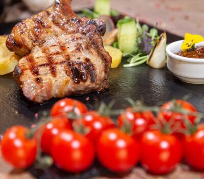 Două cotlete de porc la grătar cu roșii cherry, cartofi și sos pe farfurie neagră.