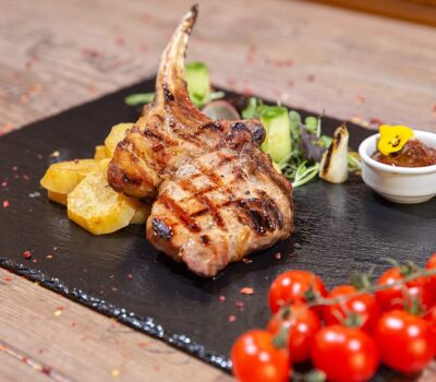 Două cotlete de miel, cartofi, salată, sos și roșii cherry pe o farfurie de ardezie.