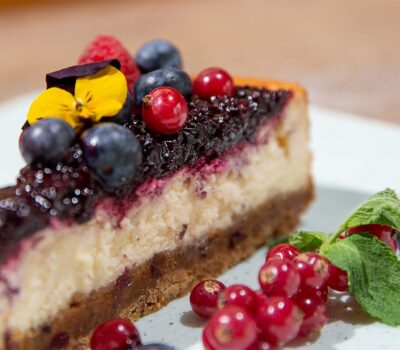 Felie de cheesecake cu fructe de pădure, afine, zmeură, coacăze și panseluțe pe o farfurie.