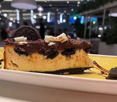 Felie de cheesecake Oreo pe farfurie galbenă, decorată cu ciocolată și biscuiți.