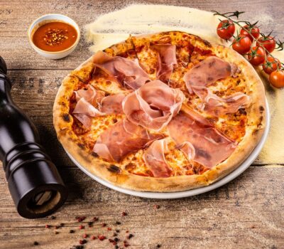 O pizza cu șuncă prosciutto lângă o râșniță de piper neagră, roșii și un sos roșu pe un fundal de lemn.