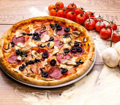 O pizza proaspătă cu pepperoni, măsline și ciuperci pe un blat alb. Roșii, ciuperci, faină pe o suprafață de lemn.