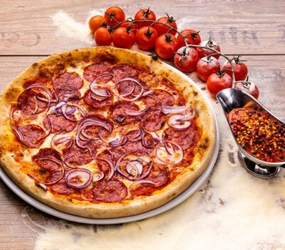 O pizza cu pepperoni și ceapă roșie, cu sos picant, roșii cherry și blat de lemn.