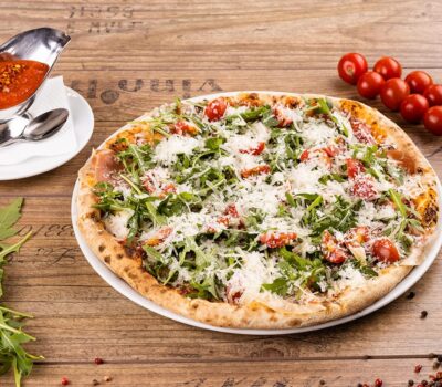 O pizza cu rucola, roșii și parmezan pe o farfurie albă cu sos.