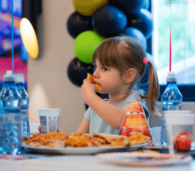 Fetiță cu pizza la o petrecere. Baloane în fundal. Băuturi și pălării tematice "Cars".