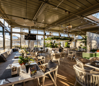 Interiorul unui restaurant luminos, cu mese, scaune, plante și lumini decorative. Ferestre mari.